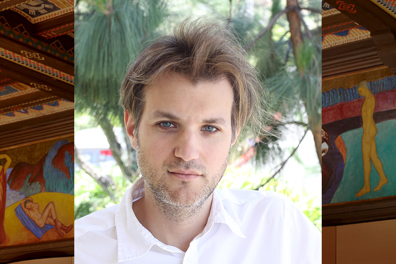 A color photograph portrait of pianist Tellef Johnson, who looks directly into the camera wearing a white collared shirt with a color photo the concert hall frescoes framing. Photo courtesy of the artist.