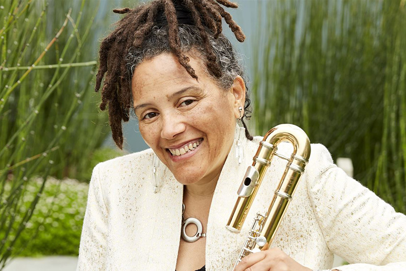 Composer and Improviser Nicole Mitchell smiles broadly into the camera, dressed in a cream and gold flecked blazer while holder a flute and seated before a garden of green equisetum. Photo by Amanda E Friedman.